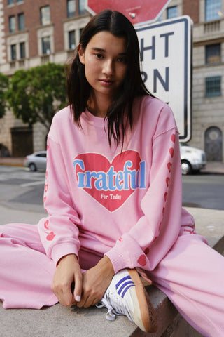 Person wearing a pink sweatshirt with 'Grateful For Today' text and hearts, sitting on a street corner.
