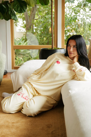 Woman in a buttercream-colored outfit with text sitting on a couch in a room with large windows and greenery.